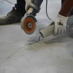 Un trabajador con guantes usa una amoladora para reparar una grieta en una solera de hormigón, mientras una manguera de aspiración recoge el polvo.