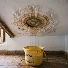 Goteo en el techo y un cubo amarillo recogen agua. Urge reparar tejado para evitar daños mayores.