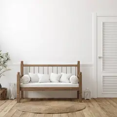 Un banco de madera rústico con cojines blancos, junto a una planta y una puerta blanca. Ideal para aprender como hacer un banco de madera.