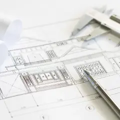 Plano arquitectónico con un **muro de carga** detallado. Se observan rollos de papel, un calibrador y un bolígrafo, listos para el diseño.