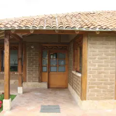 Fachada de casa con paredes de barro cocido, techo de tejas y detalles en madera. Flores rosadas y rojas adornan la entrada.