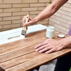 Persona aplica pintura blanca a una mesa de madera ya pintada. El proceso de como pintar madera ya pintada es sencillo con esta técnica.