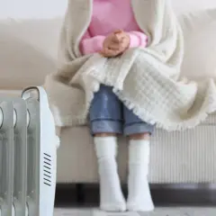 Persona abrigada con manta y calcetines sentada en sofá, junto a un radiador eléctrico, solución de calefacción sin instalación.
