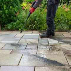 Persona con guantes rojos usa hidrolimpiadora para limpiar suelo de terraza exterior con baldosas de piedra.