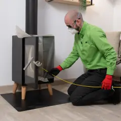 Hombre con mascarilla y guantes rojos realiza la tarea de deshollinar chimenea, usando una herramienta para limpiar el interior.