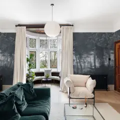 Un salón con papel pintado de bosque oscuro, un sofá verde, sillón beige y chimenea de madera. Ideas para cómo empapelar una pared con estilo.