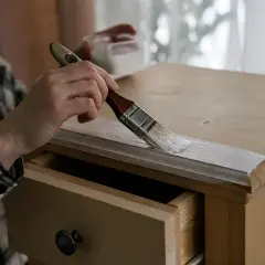 Persona pinta mesa de madera con brocha blanca. Se ve un cajón abierto.