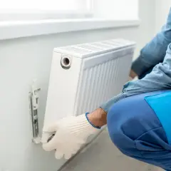 Man con guantes blancos ajusta un radiador blanco en la pared. Se está llevando a cabo la tarea de cambiar radiador de sitio.