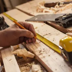 Mano marcando madera con lápiz y cinta métrica, lista para empezar a hacer una estantería de madera. Herramientas de carpintería esparcidas.