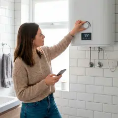 Mujer ajustando el termostato de un termo eléctrico mientras revisa su móvil. Mantenimiento termo eléctrico sencillo y moderno.