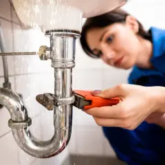 Mujer fontanera usando llave inglesa para reparar tubería con fugas bajo el lavabo.
