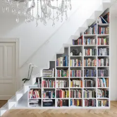 Estantería blanca de pladur para cerrar hueco bajo escalera, llena de libros y con una lámpara de araña.
