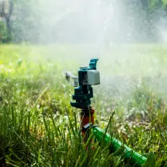 Aspersor de instalación de riego en acción, rociando agua sobre césped verde y frondoso bajo la luz del sol.