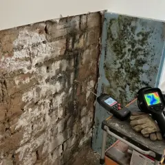 Herramientas para diagnosticar y como aislar una pared de la humedad: medidor de humedad, cámara térmica y guantes sobre una pared de ladrillo con moho.