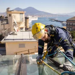 Trabajador con arnés realiza reparación tejado sin quitar tejas, con vistas a la bahía y el Vesubio.