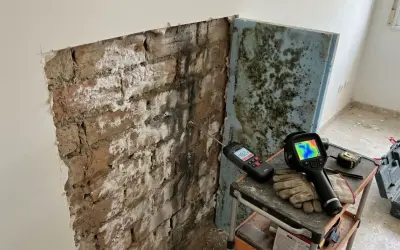 Herramientas para diagnosticar y como aislar una pared de la humedad: medidor de humedad, cámara térmica y guantes sobre una pared de ladrillo con moho.
