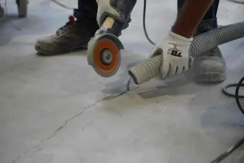 Un trabajador con guantes usa una amoladora para reparar una grieta en una solera de hormigón, mientras una manguera de aspiración recoge el polvo.
