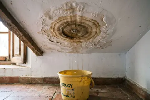 Goteo en el techo y un cubo amarillo recogen agua. Urge reparar tejado para evitar daños mayores.