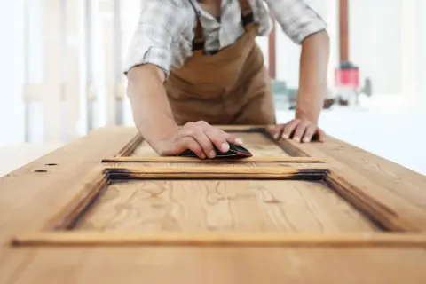 Carpintero lijando una puerta de sapelly para modernizarla, revelando la veta de la madera.
