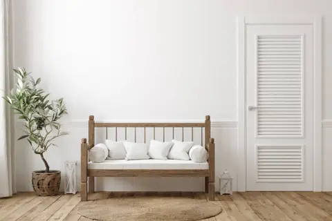 Un banco de madera rústico con cojines blancos, junto a una planta y una puerta blanca. Ideal para aprender como hacer un banco de madera.
