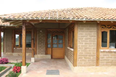 Fachada de casa con paredes de barro cocido, techo de tejas y detalles en madera. Flores rosadas y rojas adornan la entrada.