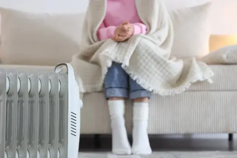 Persona abrigada con manta y calcetines sentada en sofá, junto a un radiador eléctrico, solución de calefacción sin instalación.