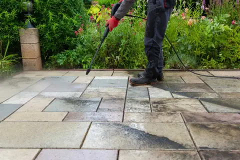 Persona con guantes rojos usa hidrolimpiadora para limpiar suelo de terraza exterior con baldosas de piedra.