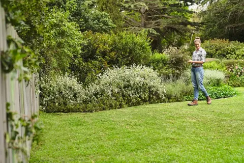 Mujer sonríe en un jardín exuberante, con un seto de arbustos blancos y verdes, y un gran chopo al fondo.