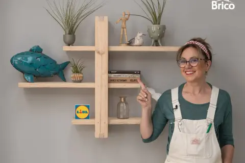 Estantería DIY de madera con pez decorativo, plantas y libros. Una mujer sonríe y señala la estantería.