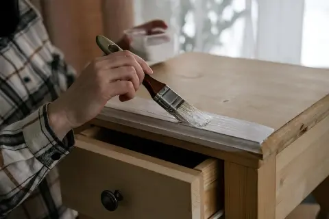 Persona pinta mesa de madera con brocha blanca. Se ve un cajón abierto.