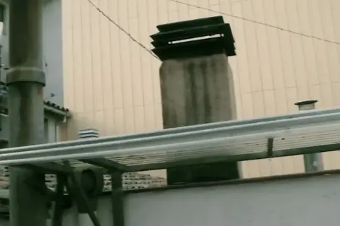 La chimenea del vecino desprende humo que me entra. Se ven varias chimeneas y tuberías en un tejado.