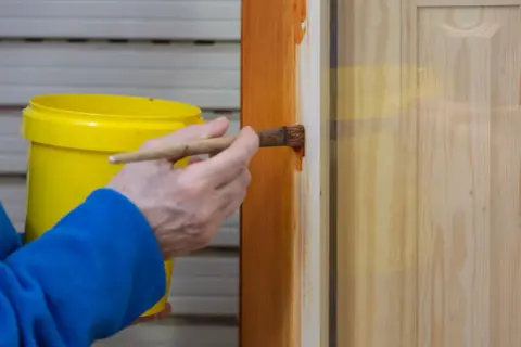 Persona aplica barniz a una puerta de madera con un pincel. El proceso de lacar puertas protege y embellece la madera.