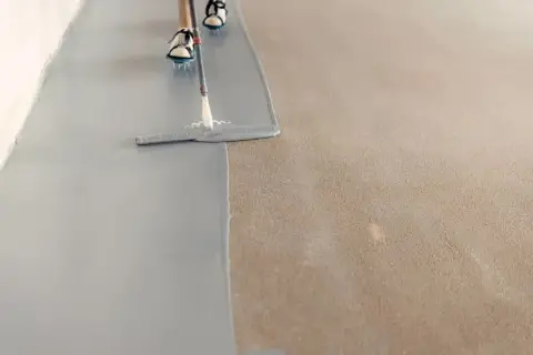 Persona pintando el suelo con una llana, aplicando una capa gris sobre una superficie texturizada.