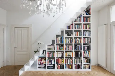 Estantería blanca de pladur para cerrar hueco bajo escalera, llena de libros y con una lámpara de araña.