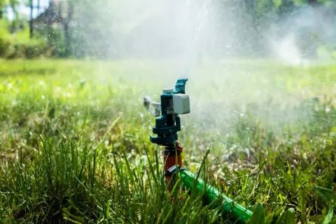 Aspersor de instalación de riego en acción, rociando agua sobre césped verde y frondoso bajo la luz del sol.