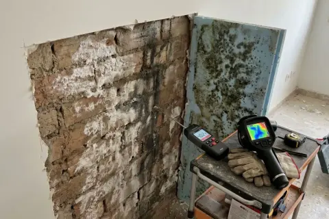 Herramientas para diagnosticar y como aislar una pared de la humedad: medidor de humedad, cámara térmica y guantes sobre una pared de ladrillo con moho.