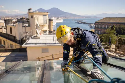 Trabajador con arnés realiza reparación tejado sin quitar tejas, con vistas a la bahía y el Vesubio.
