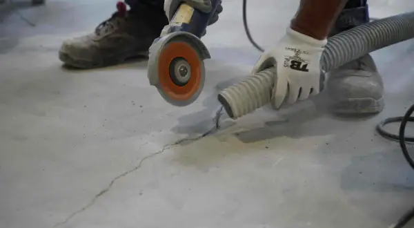 Un trabajador con guantes usa una amoladora para reparar una grieta en una solera de hormigón, mientras una manguera de aspiración recoge el polvo.