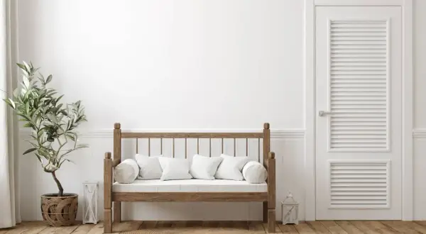 Un banco de madera rústico con cojines blancos, junto a una planta y una puerta blanca. Ideal para aprender como hacer un banco de madera.