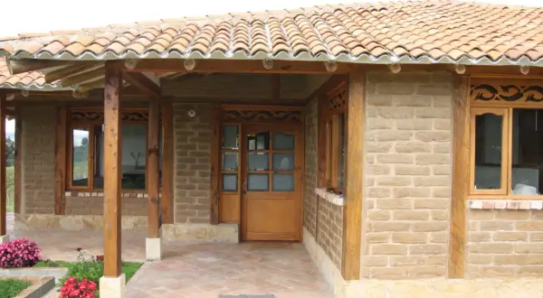Fachada de casa con paredes de barro cocido, techo de tejas y detalles en madera. Flores rosadas y rojas adornan la entrada.
