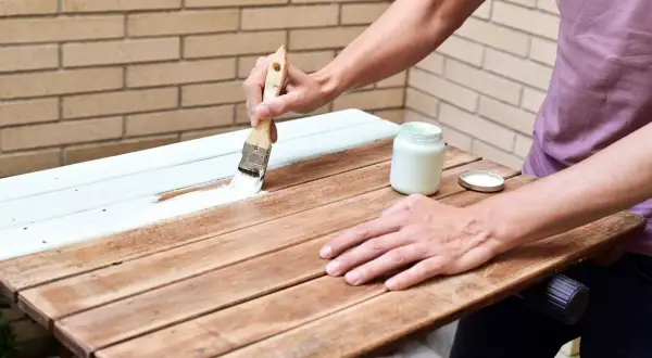 Persona aplica pintura blanca a una mesa de madera ya pintada. El proceso de como pintar madera ya pintada es sencillo con esta técnica.