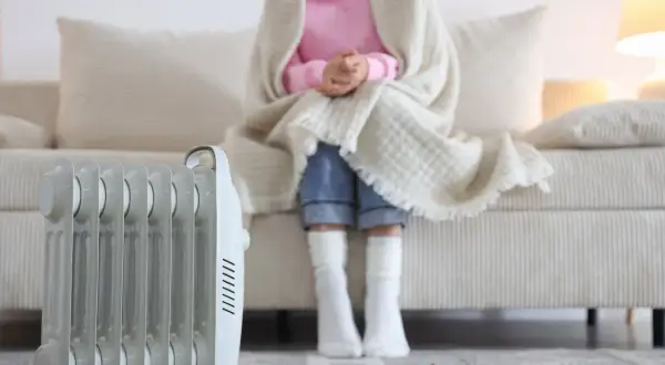 Persona abrigada con manta y calcetines sentada en sofá, junto a un radiador eléctrico, solución de calefacción sin instalación.