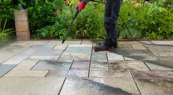 Persona con guantes rojos usa hidrolimpiadora para limpiar suelo de terraza exterior con baldosas de piedra.