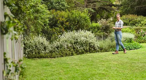 Mujer sonríe en un jardín exuberante, con un seto de arbustos blancos y verdes, y un gran chopo al fondo.