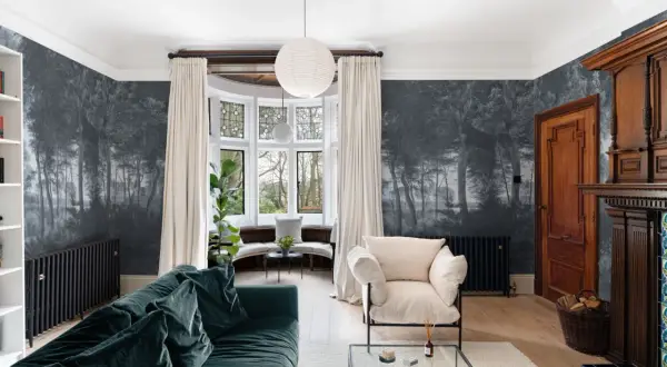 Un salón con papel pintado de bosque oscuro, un sofá verde, sillón beige y chimenea de madera. Ideas para cómo empapelar una pared con estilo.