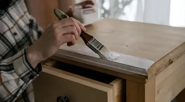 Persona pinta mesa de madera con brocha blanca. Se ve un cajón abierto.