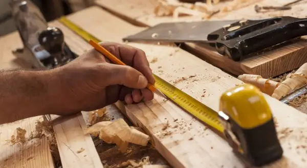 Mano marcando madera con lápiz y cinta métrica, lista para empezar a hacer una estantería de madera. Herramientas de carpintería esparcidas.