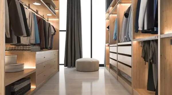 Un vestidor cerrado de madera clara, con ropa colgada y cajones organizados. Un puf beige y cortinas oscuras completan la escena.