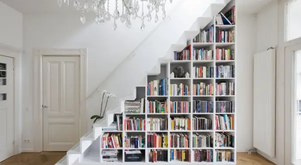 Estantería blanca de pladur para cerrar hueco bajo escalera, llena de libros y con una lámpara de araña.