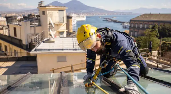 Trabajador con arnés realiza reparación tejado sin quitar tejas, con vistas a la bahía y el Vesubio.